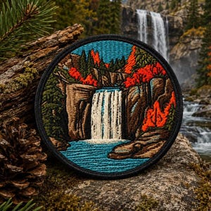 May include: A circular embroidered patch featuring a landscape scene of a waterfall cascading over brown rocky cliffs. The design includes vibrant orange and green trees, a deep blue pool at the base, and a thick black stitched border edge.