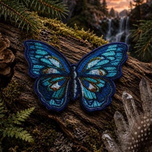 May include: An embroidered butterfly patch featuring shades of deep navy, bright cyan, and white thread. The wings display intricate symmetrical patterns with dark outlines and subtle brown accents, creating a detailed insect design.