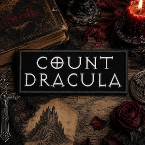 May include: A rectangular black embroidered patch featuring the white text "COUNT DRACULA" in a serif font, with a cross symbol replacing the letter "O" in the word "COUNT". The patch has a clean, stitched border and a flat, fabric surface.