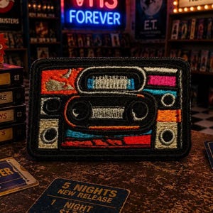 May include: Embroidered patch featuring a retro cassette tape design with orange, blue, and pink accents. The patch is rectangular with a black border. The background includes VHS tapes, popcorn, and a neon sign that says "VHS FOREVER".