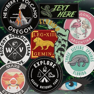 May include: A collection of embroidered patches featuring various designs, including a volcano, a snake, a lion, a manatee, flamingos, and a tent. The patches are all circular and have black borders. The text on the patches includes "Oregon", "Florida", "Three Sisters Springs", "Gemina", "Explore Acadia National Park", and "Text Here".
