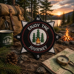 May include: A black star-shaped embroidered patch featuring a white center with three green pine trees and a small campfire. The text ENJOY THE JOURNEY is stitched in white around the center. The patch has white compass points labeled N, S, E, W.