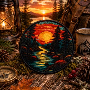 Può includere: Una toppa ricamata circolare che raffigura un vivace tramonto su un fiume tortuoso e una foresta di pini. Il design utilizza fili arancioni, gialli, rossi e blu scuro per ritrarre un paesaggio con un bordo nero.