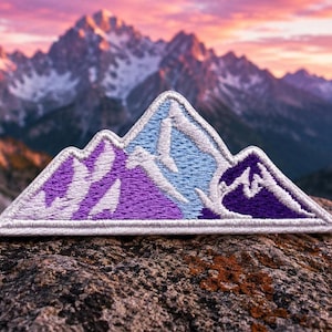 May include: A triangular embroidered patch featuring a stylized mountain range design. The patch uses shades of light blue, medium purple, and dark violet thread with white stitching outlining the peaks and the base of the mountain shape.