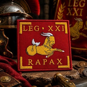Può includere: Una toppa quadrata rossa con la scritta "LEG XXI RAPAX" in giallo. La toppa presenta una creatura gialla simile a una capra. Si tratta probabilmente di un emblema militare o storico, forse un'insegna della legione romana.