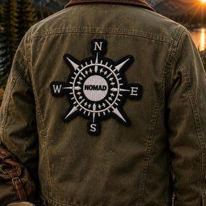 May include: An olive green jacket featuring a large, circular embroidered patch on the back. The patch displays a black and white compass design with the word NOMAD in the center and the letters N, S, E, and W positioned at the cardinal points.