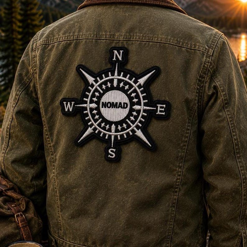May include: An olive green jacket featuring a large, circular embroidered patch on the back. The patch displays a black and white compass design with the word NOMAD in the center and the letters N, S, E, and W positioned at the cardinal points.