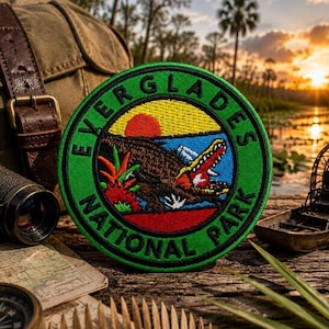 May include: A round green Everglades National Park patch featuring a brown alligator with an open mouth, a red sun, and the text "EVERGLADES NATIONAL PARK". The patch is surrounded by a green border and is set against a backdrop of a sunset over a lake.
