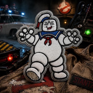 May include: Embroidered patch featuring the Stay Puft Marshmallow Man from Ghostbusters. The patch is white with a blue sailor collar, red neckerchief, and black outline. The background includes Ghostbusters-themed props and a vehicle.