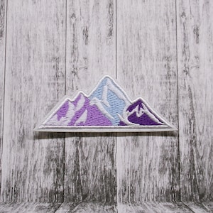 Könnte beinhalten: Lila und weiß gestickter Berg-Patch. Der Patch ist ein schlichtes Design mit drei Bergen mit weißem Umriss.
