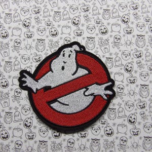 Puede incluir: Un parche bordado rojo y blanco con un fantasma en forma del logotipo de los Cazafantasmas. El fantasma es blanco con un contorno plateado y está dentro de un símbolo 'no'.