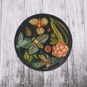 May include: A round black patch with an embroidered design of moths, butterflies, and flowers on a dark background. The design features a variety of colors, including green, yellow, orange, and pink.