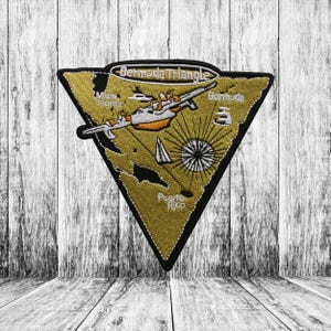 Op de afbeelding: Geborduurde patch in een driehoekige vorm met een zwarte rand. De patch toont een kaart van de Bermuda Driehoek met een vliegtuig, kompas en de tekst "Bermuda Triangle", "Miami Florida", "Bermuda" en "Puerto Rico".