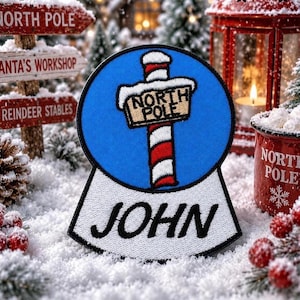 Pode incluir: Patch bordado com um letreiro do Pólo Norte sobre um fundo azul, com o nome "JOHN" abaixo. O letreiro está num poste listrado de vermelho e branco. O patch está inserido numa cena de neve com decorações de Natal.