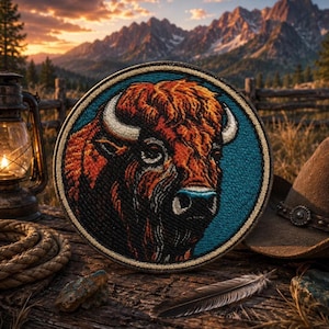 May include: A circular embroidered patch featuring a detailed portrait of a brown bison with curved horns against a solid teal background. The patch has a thick, light-coloured stitched border and displays intricate thread work in shades of orange, brown, and black to define the animal's fur and facial features.