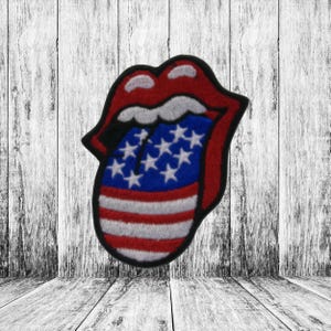 May include: Embroidered patch featuring the iconic Rolling Stones tongue and lips logo. The tongue is designed with the American flag, showcasing red and white stripes and blue with white stars. The patch has a black border and is set against a wood background.