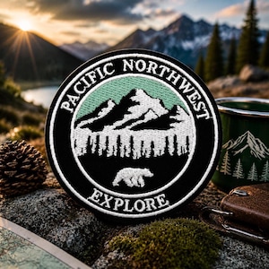 Może przedstawiać: Okrągła czarna naszywka z białymi napisami "PACIFIC NORTHWEST" i "EXPLORE". Naszywka przedstawia pasmo górskie, drzewa i niedźwiedzia. Widoczny jest zielony kubek emaliowany z motywem gór i drzew.