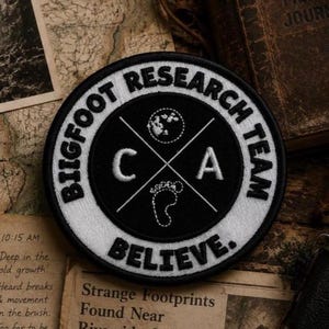 May include: A circular embroidered patch with a black center and white border. The text reads BIGFOOT RESEARCH TEAM and BELIEVE. The center features a white moon, a footprint, and the letters C and A separated by two intersecting diagonal lines.