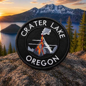 Może przedstawiać: Okrągła czarna naszywka z białym napisem "CRATER LAKE OREGON". Naszywka przedstawia haftowany obraz wulkanu w erupcji z pomarańczowymi i szarymi detalami. Tło przedstawia jezioro, góry i zachód słońca.