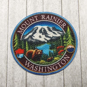 May include: A round, blue and brown embroidered patch with the text "Mount Rainier Washington" and a depiction of a mountain, a lake, trees, and a bear.