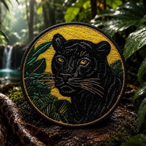 May include: A circular embroidered patch featuring a black panther with yellow eyes, set against a vibrant yellow background with green tropical foliage. The design is framed by a black stitched border, showcasing detailed thread work.