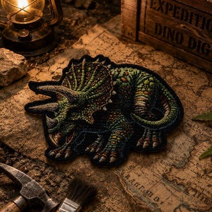 Könnte beinhalten: Ein bestickter Aufnäher mit einem detaillierten Triceratops-Dinosaurier in Grün-, Braun- und Gelbtönen. Der Dinosaurier ist in einer kauernden Position dargestellt, wobei seine drei Hörner und der Nackenschild durch feine Stickereien deutlich erkennbar sind.