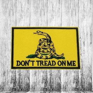 Può includere: Una toppa gialla con bordo nero e un serpente avvolto con testa verde. Il testo "Don't Tread On Me" è stampato in nero sotto il serpente.