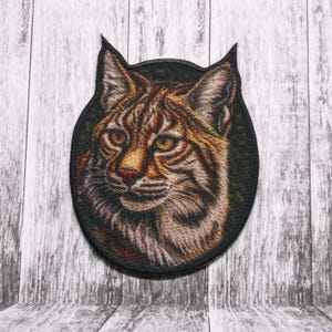 May include: Close-up of a brown and tan lynx with yellow eyes. The lynx is looking directly at the camera. The image is a circular patch with a black border.