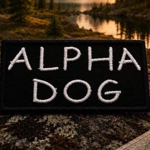 May include: A rectangular black patch with white embroidered text that reads ALPHA DOG in two lines. The patch features a clean, stitched border and is presented with a clear, high-contrast design suitable for attachment to various fabric items.