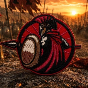 May include: A circular embroidered patch featuring a Spartan warrior in a black helmet and red cape, holding a round shield and a spear. The design is set against a red background with radiating lines, framed by a black border with a spear tip.