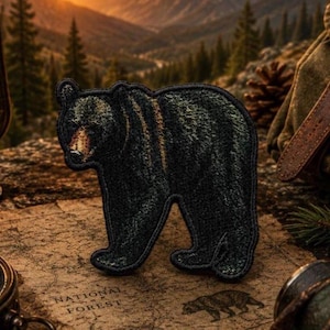 May include: Embroidered patch of a black bear in a walking pose. The patch has a detailed texture and is set against a backdrop of a map with "NATIONAL FOREST" printed on it. The bear patch is a camping or outdoor theme.