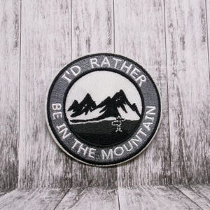 Peut inclure: Un écusson brodé gris et blanc avec le texte "I'd Rather Be In The Mountain" et une silhouette noire de montagnes.
