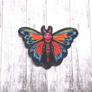 May include: A colorful butterfly patch with red, orange, yellow, blue, and pink wings. The butterfly has a black body with a pink head and a skeletal design.