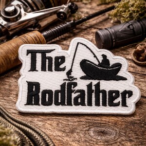 May include: A white embroidered patch with a black border featuring the text The Rodfather in a bold font. To the right of the text is a black silhouette graphic of a person sitting in a small boat holding a fishing rod with a line in the water.