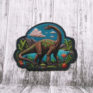 May include: Embroidered patch depicting a brown dinosaur with a long neck standing in a lush green jungle setting. The dinosaur is surrounded by green plants and flowers, and the background is a blue sky with white clouds.