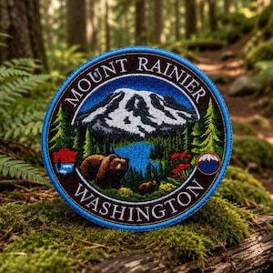 May include: A circular embroidered patch featuring a snow-capped mountain, a blue lake, green pine trees, and two brown bears. The text MOUNT RAINIER WASHINGTON is stitched in white along the border. The patch has a bright blue outer edge.