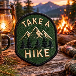 May include: Green embroidered patch with the words "TAKE A HIKE" and a mountain and tree design. The patch is on a wooden surface, with a lantern, mug, and hiking boots in the background. The patch is approximately 3 inches wide.
