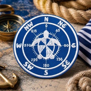 May include: A round, blue embroidered patch featuring a white compass rose design with a sea turtle at the center. The patch includes directional markings and is surrounded by a white border. Nautical theme.