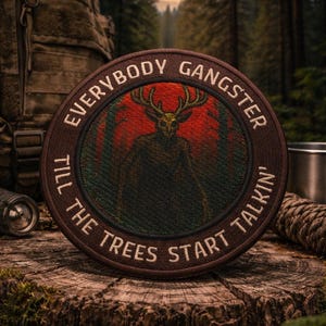 May include: A circular brown patch with the text "EVERYBODY GANGSTER TILL THE TREES START TALKIN'". The patch depicts a deer-like figure with antlers against a red and green forest backdrop. The patch is on a wooden surface.