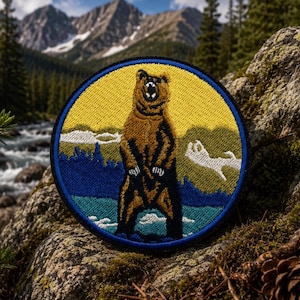 May include: A circular embroidered patch featuring a brown bear standing on its hind legs in the center. The background depicts a yellow sky, green forest, and blue water. The patch has a thick dark blue border and intricate thread detailing.