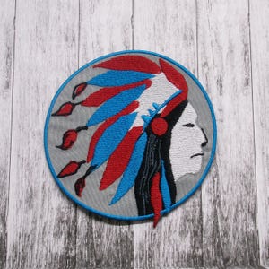 Puede incluir: Un parche redondo bordado que representa a una persona nativa americana de perfil con un tocado de plumas rojas, blancas y azules. El fondo es gris.