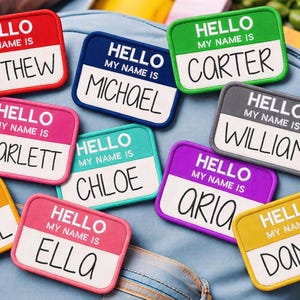Op de afbeelding: Een verzameling kleurrijke naamlabels met de tekst "HELLO MY NAME IS" en verschillende namen zoals Matthew, Michael, Carter, Scarlett, Chloe, William, Aria, Abigail, Ella en Daniel. De labels zijn in verschillende kleuren.