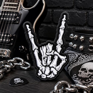 May include: Embroidered patch featuring a white skeleton hand making the rock and roll gesture, outlined in black. The patch is surrounded by a black electric guitar, a studded wristband, a skull bandana, a guitar pick with a skull and crossbones, and a silver chain.