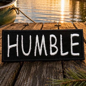 May include: A rectangular black embroidered patch featuring the word HUMBLE stitched in white capital letters. The patch has a clean, finished border and is positioned horizontally, displaying a clear contrast between the dark fabric and the white thread.
