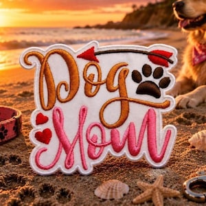 May include: A white embroidered patch featuring the text Dog Mom in brown and pink cursive script. The design includes a red arrow pointing right, a brown paw print, and two small red hearts. The patch has a scalloped edge and white border.