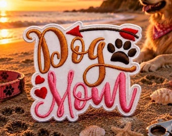 May include: A white embroidered patch featuring the text Dog Mom in brown and pink cursive script. The design includes a red arrow pointing right, a brown paw print, and two small red hearts. The patch has a scalloped edge and white border.