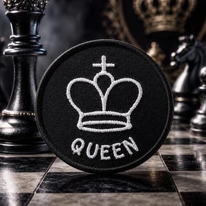Peut inclure: Un écusson rond noir avec une couronne blanche brodée et le mot "QUEEN". L'écusson est posé sur un échiquier, avec des pièces d'échecs noires en arrière-plan. Un article décoratif.