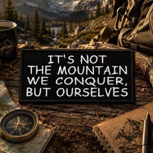 Pode incluir: Um emblema retangular preto bordado com o texto branco: IT'S NOT THE MOUNTAIN WE CONQUER, BUT OURSELVES. O emblema possui uma borda limpa e acabada, apresentando-se como um acessório de tecido plano e durável.