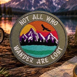 May include: A circular embroidered patch with an olive green border featuring the text NOT ALL WHO WANDER ARE LOST in white. The centre depicts a yellow sun, purple mountains with white snow caps, a black bird silhouette, and a teal lake.