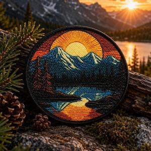 May include: A circular embroidered patch featuring a landscape scene with a bright yellow sun rising over blue mountain peaks. The design includes a dark forest, a reflective lake, and orange sky gradients, all enclosed within a black stitched border.